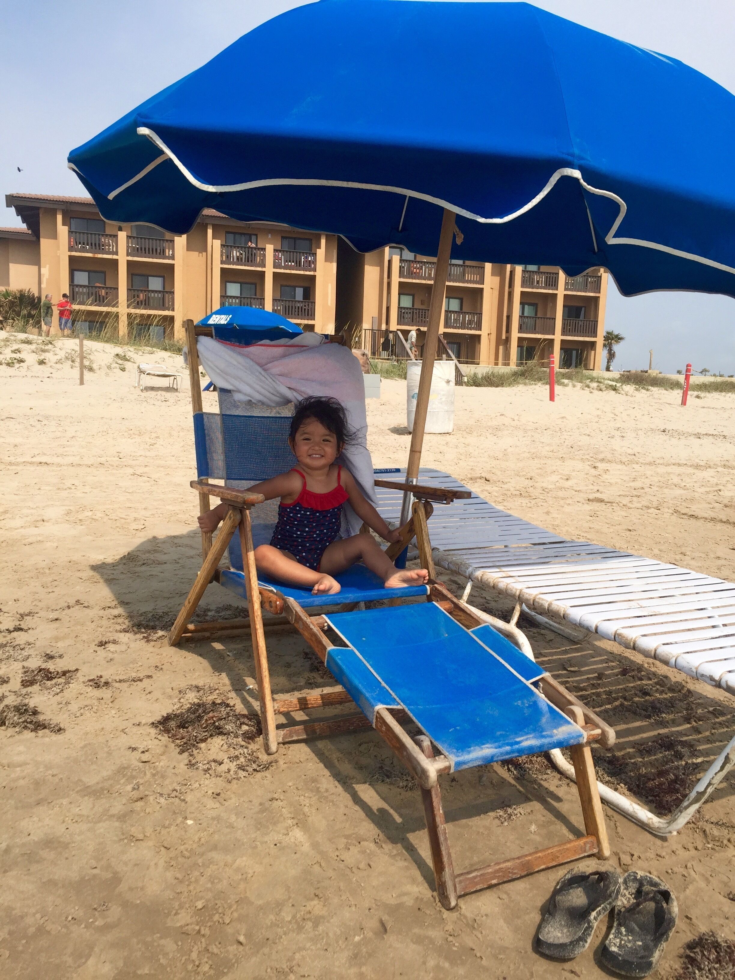 Un niño sentado bajo un paraguas azul en una silla de playa.