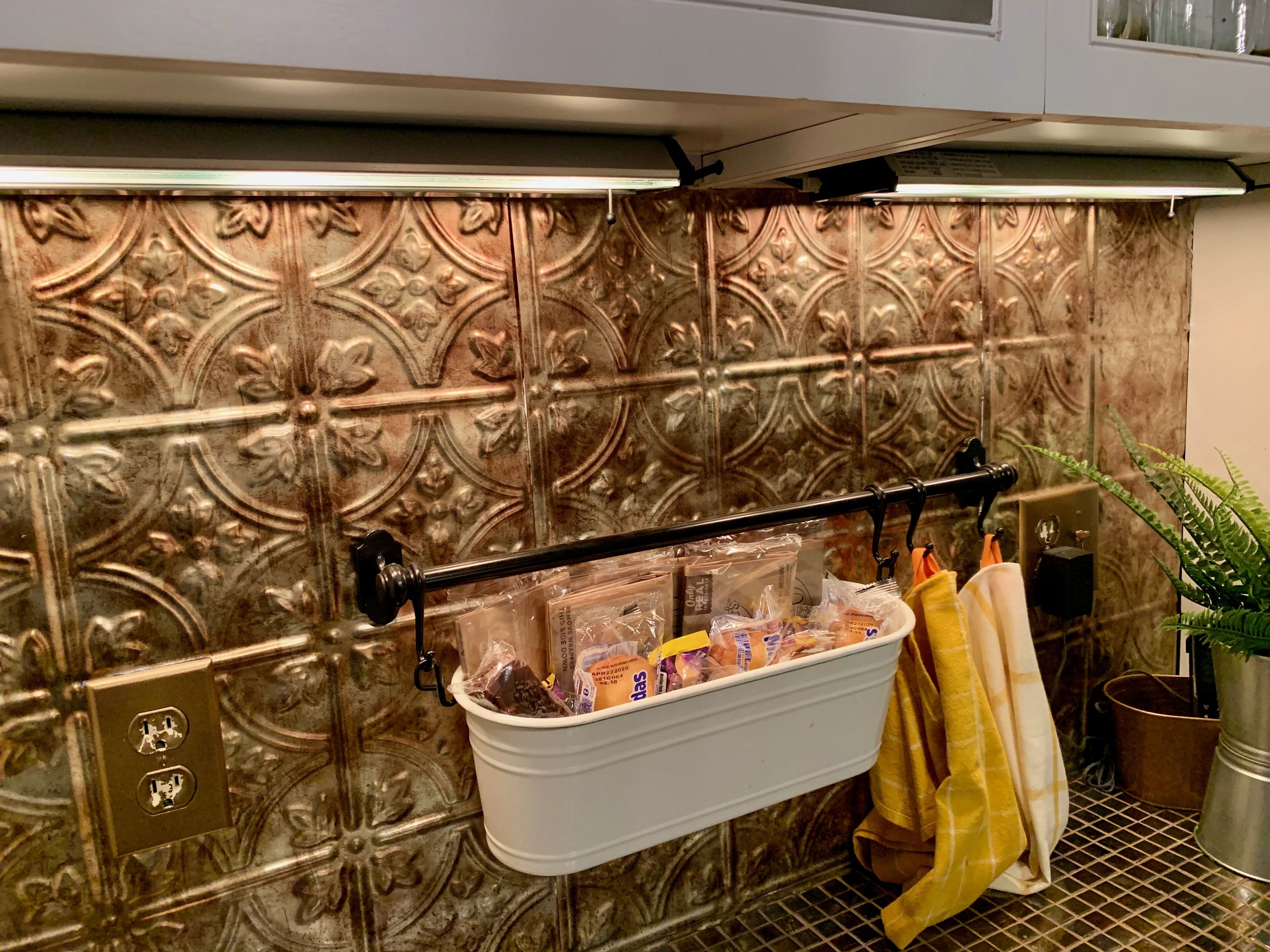 A kitchen with a decorative backsplash, a hanging rack with packaged food items, and a yellow towel on the counter.