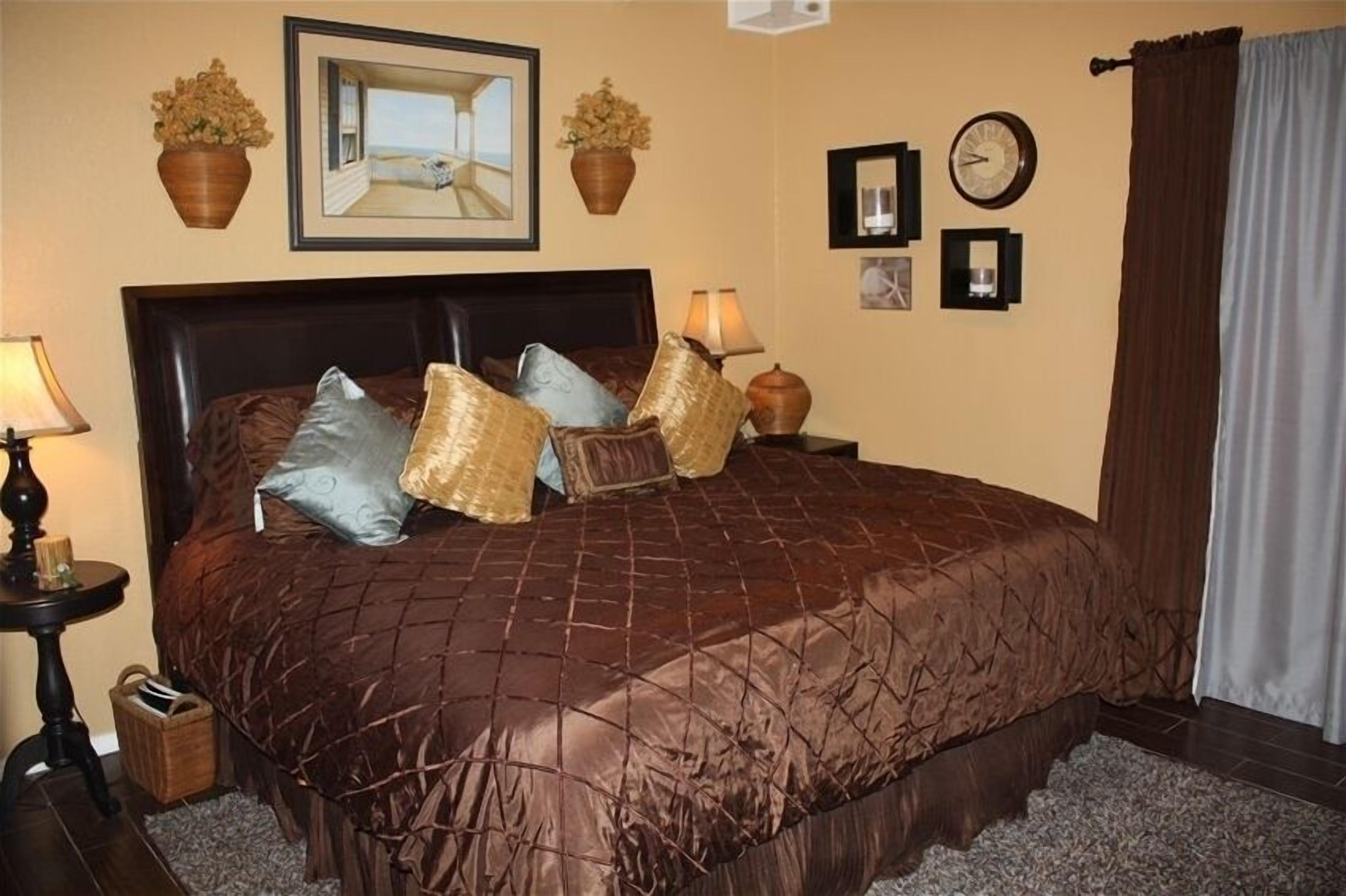 A neatly made bed with a brown quilt, two side tables with lamps, a clock, and framed pictures on the wall.