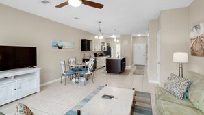 A spacious living room with a flat-screen TV, a dining area with chairs, a kitchen with cabinets, and a ceiling fan. - ⭐Amazing Location-Complex Pool-Balcony and Patio-Corner Unit-2BR Gulf Walk 606⭐ (Destin)