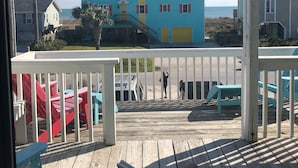 A view from a window overlooking a wooden deck with chairs, a building with a blue facade, and a clear sky. - Prime Location 2nd row!!! Beautiful beach views from the deck!!  (Emerald Isle)