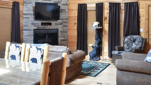 L’intérieur d’un chalet en bois est doté d’une cheminée en pierre, d’un téléviseur fixé au-dessus, d’une table à manger avec des chaises, d’un canapé et d’un ventilateur de plafond.