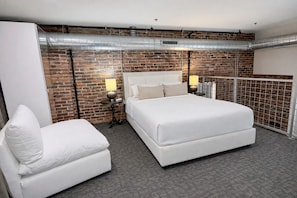 Quarto de hotel moderno com parede de tijolos, uma cama grande, uma poltrona branca e uma pequena mesa com abajur.