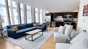 A modern living room with a sectional sofa, a coffee table, and a grey couch. A kitchen with dark cabinets and stainless steel appliances is visible in the background.