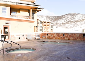 Ein Poolbereich mit zwei Whirlpools, einer Steinmauer, einer Holzterrasse und einem Gebäude mit Balkon im Hintergrund.