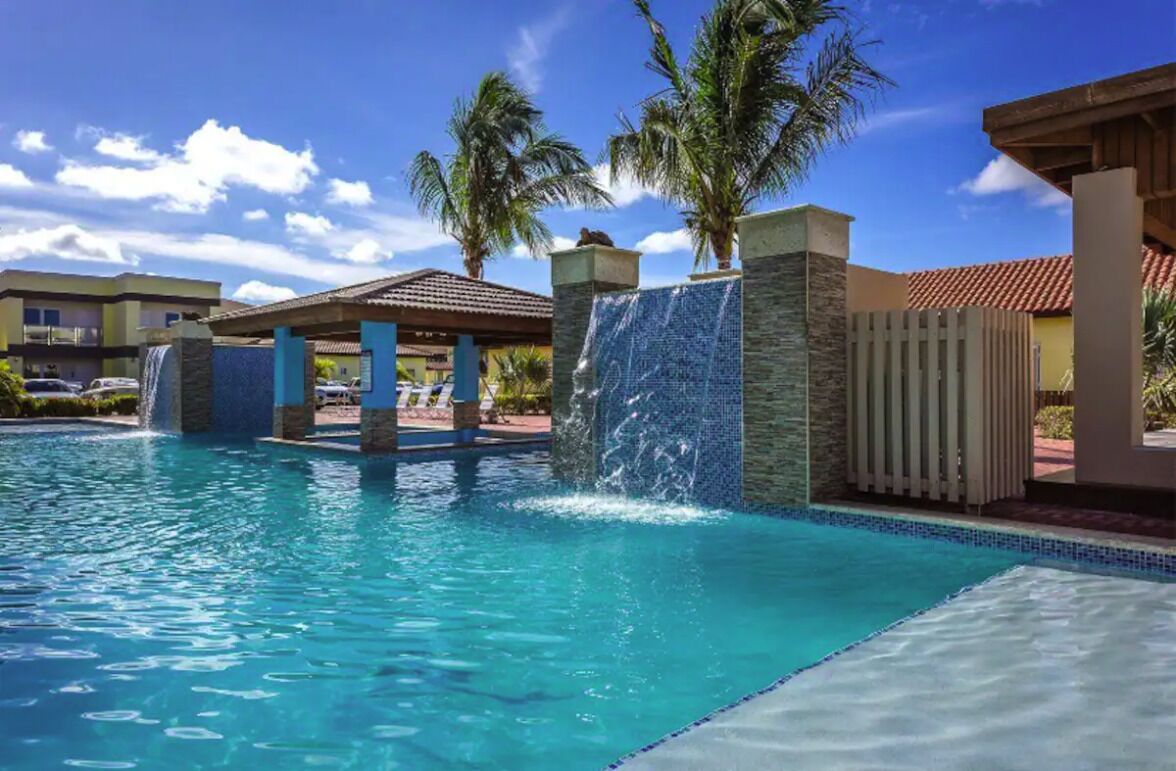 A swimming pool with a waterfall feature, palm trees, and a clear blue sky.