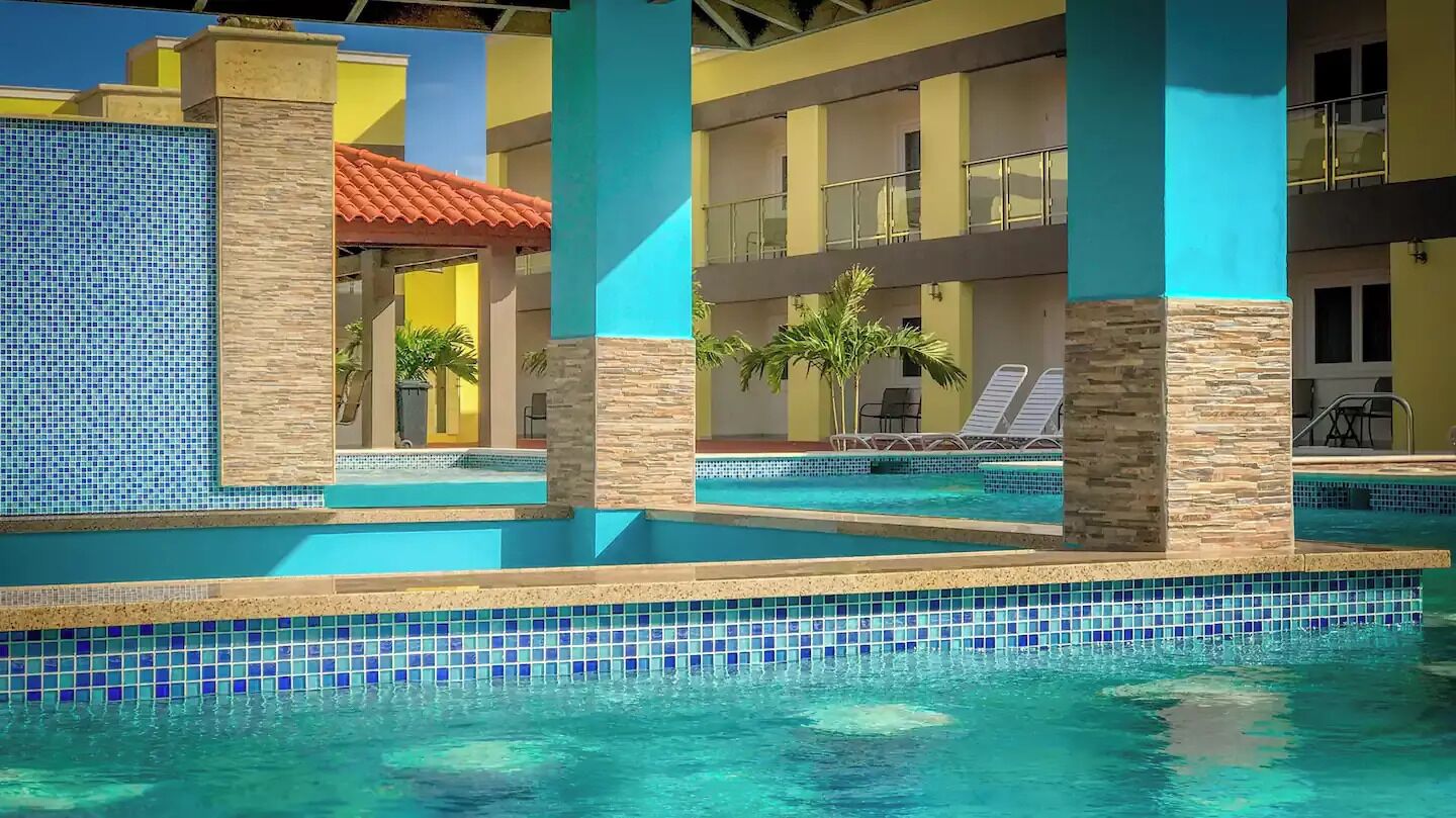 A pool area with tiled walls and a clear blue pool, surrounded by lounge chairs and palm trees.