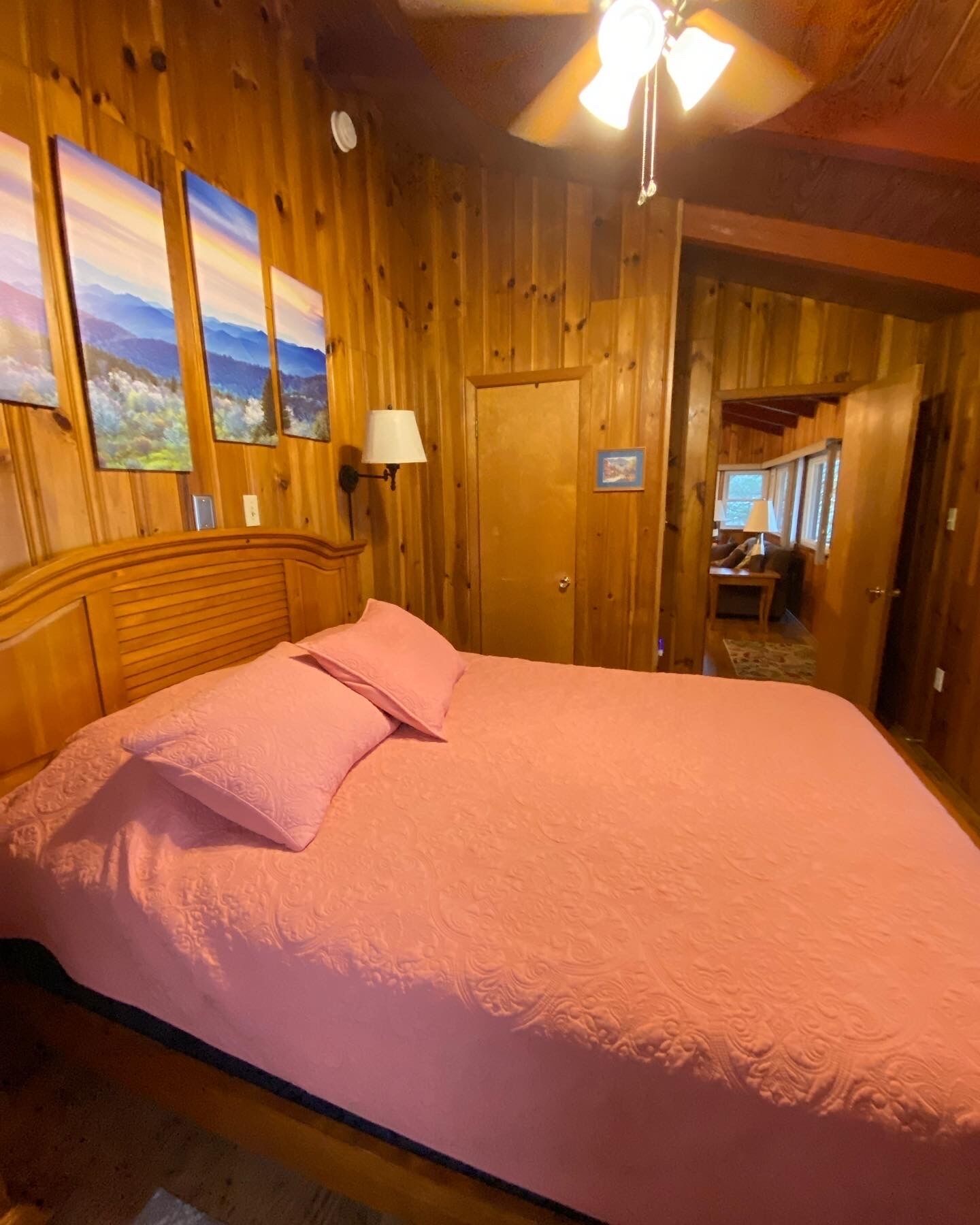 A wooden-paneled room with a bed, a ceiling fan, and a door leading to another room.
