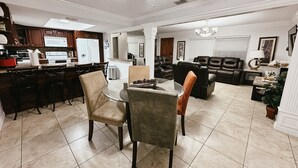 A modern kitchen with dark wooden cabinets, a white refrigerator, and a stainless steel stove. There is a dining area with a glass table and chairs, and a living room with leather sofas and a chandelier. - Affordable 3 Bedroom Home W / Private Pool. Minutes from Orlando's theme parks (Orlando)