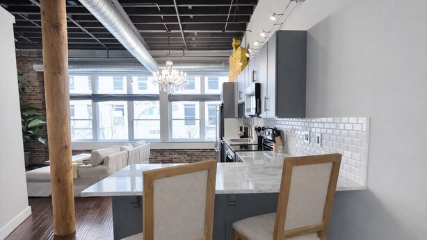 A modern kitchen with a large island, two wooden chairs, and a view of the city through large windows. - Downtown Music City Loft living! ~Steps to the Ryman and Broadway. (Nashville)