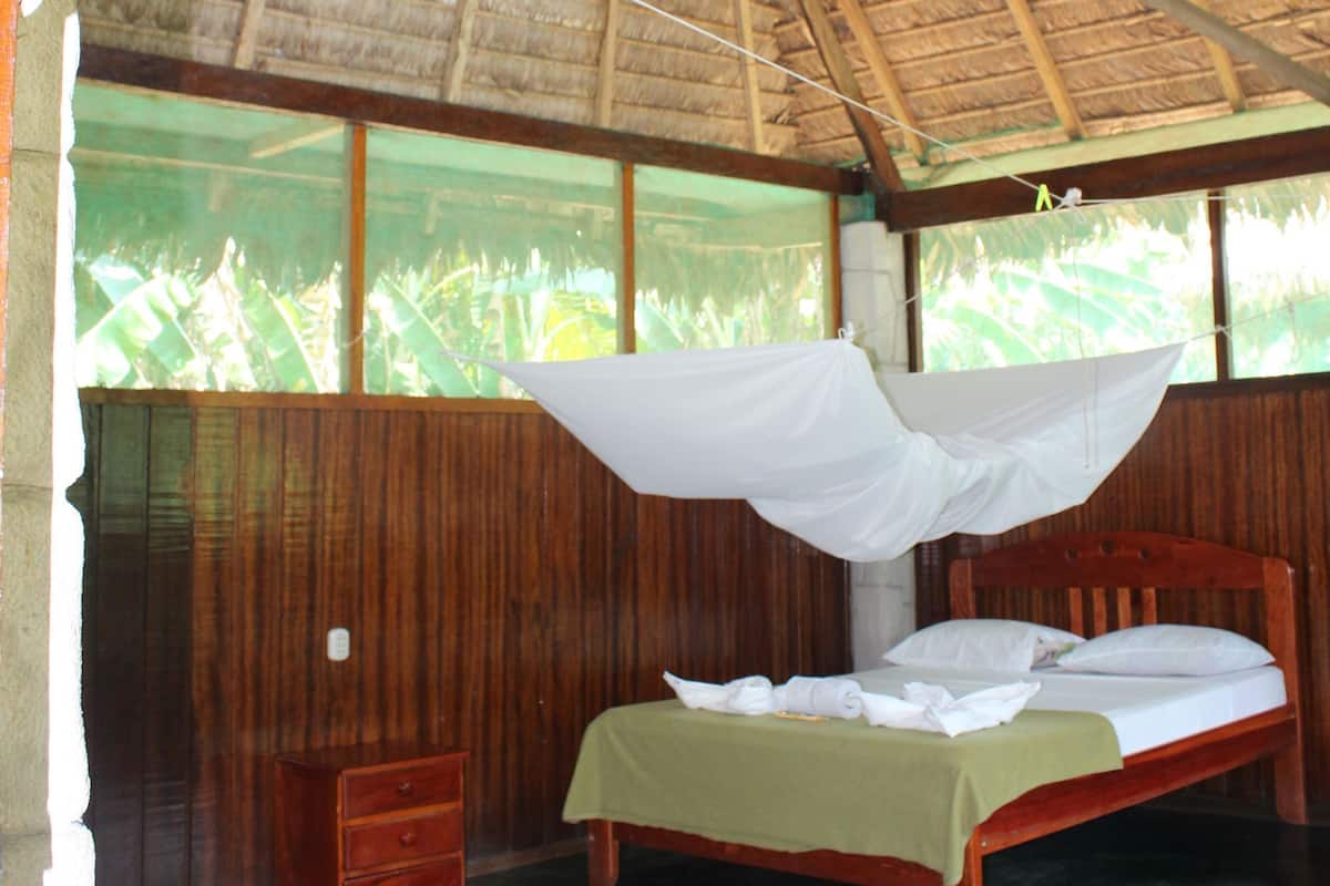 A room with a bed, wooden walls, a thatched roof, and large windows.