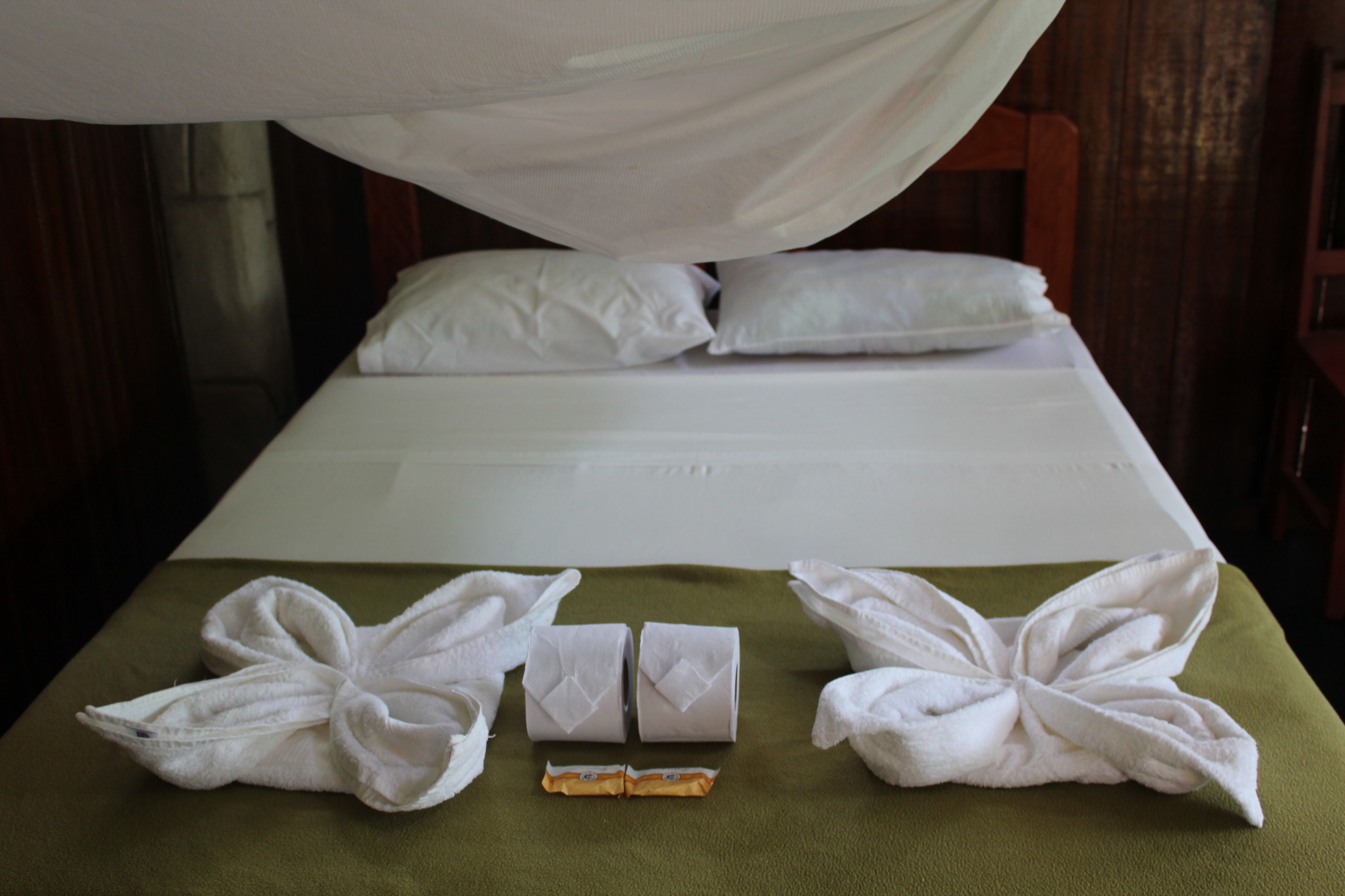 Una cama bien tendida con sábanas y almohadas blancas, una colcha verde y dos toallas dobladas sobre la cama.