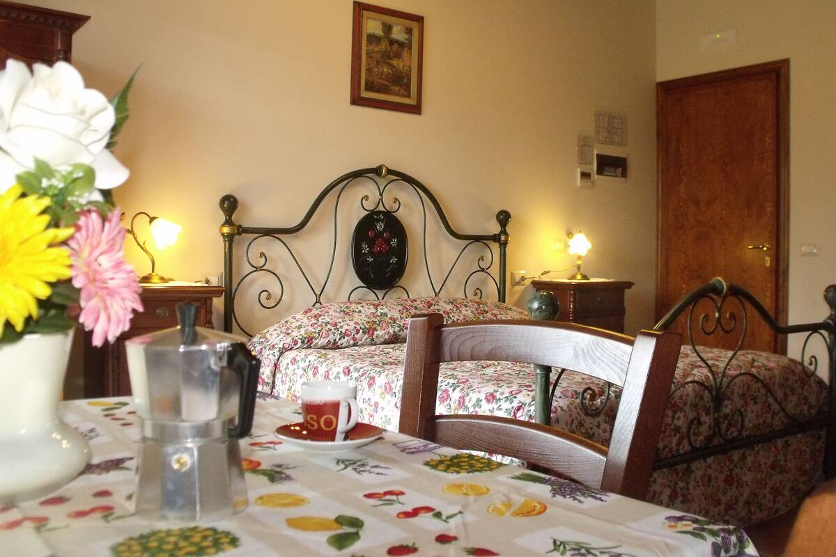 A bedroom with a floral bedspread, a bedside table with a lamp, and a vase with flowers.