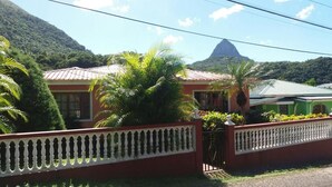 A house with a red exterior and a white fence, surrounded by lush greenery and a mountain in the background.