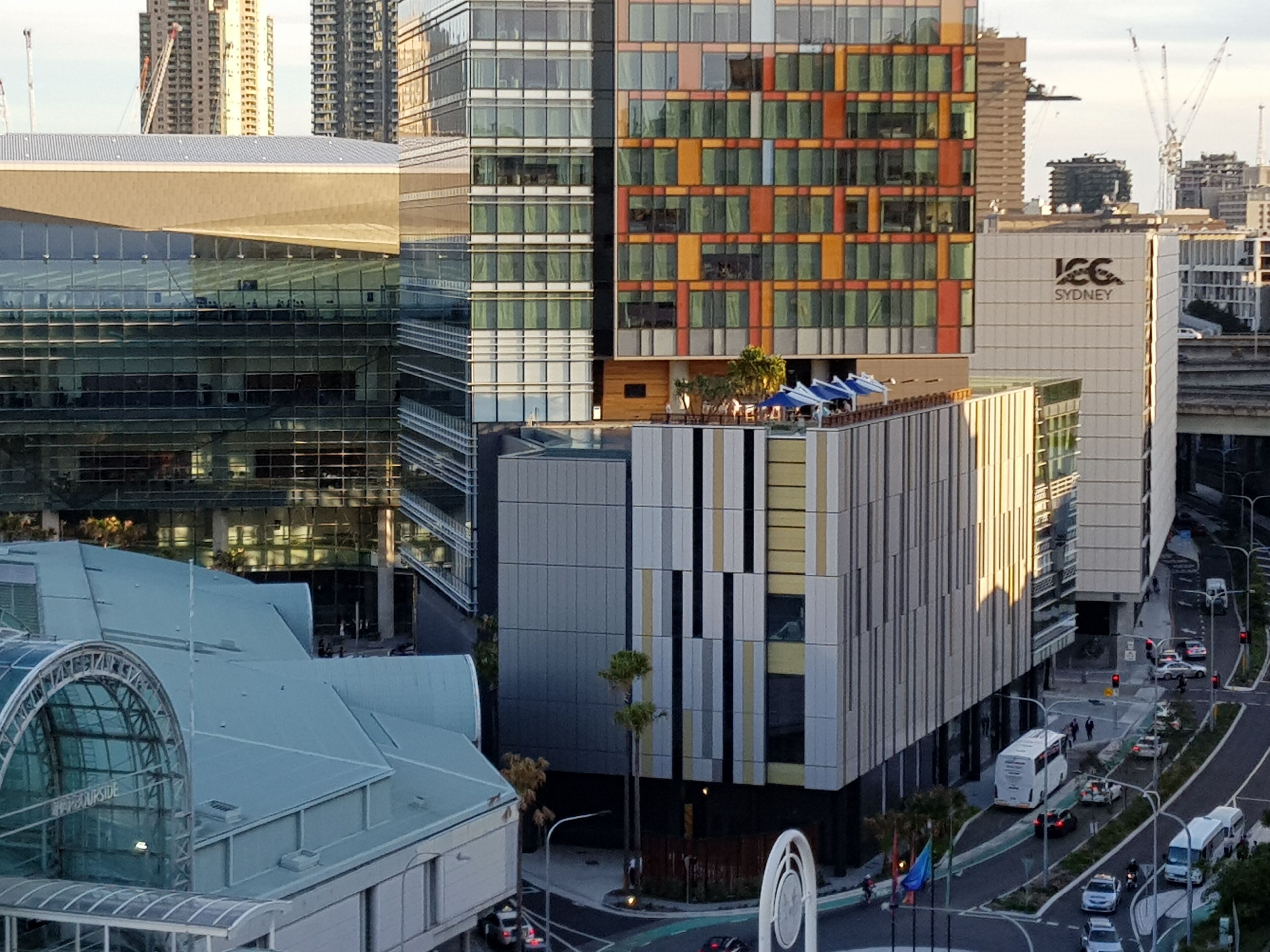 Um edifício moderno urbano com fachada de vidro, logotipo em destaque e vista da rua com veículos e faixa de pedestres.