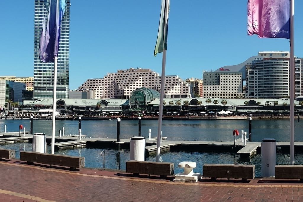 A waterfront marina with docked boats, a marina building, and a city skyline in the background.