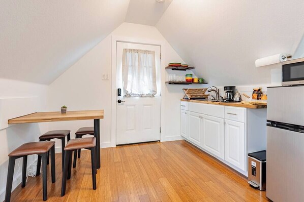 A compact kitchen with a wooden table, four stools, white cabinets, a microwave, and a coffee machine.