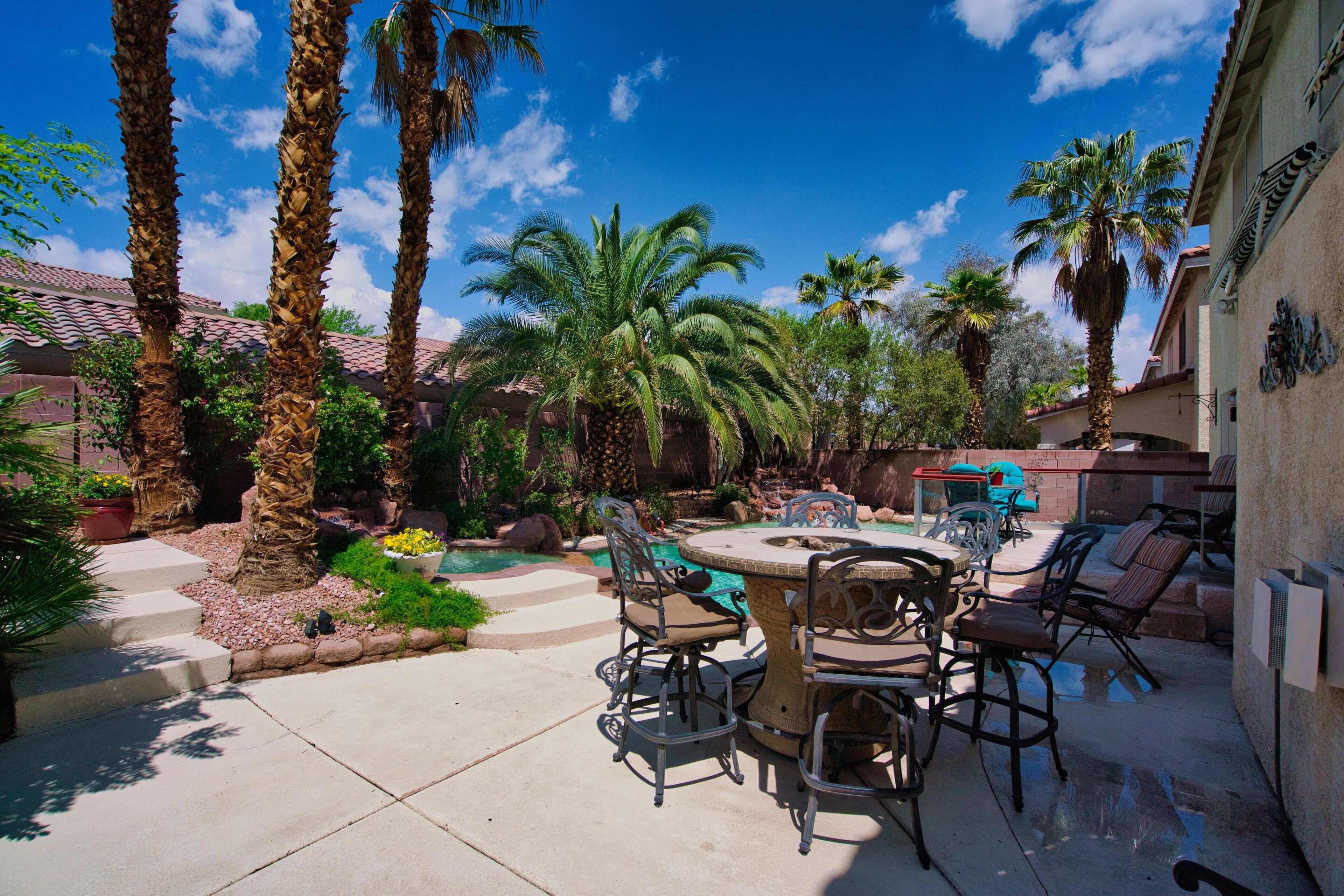 An outdoor patio with a round table and chairs, surrounded by palm trees and a swimming pool.