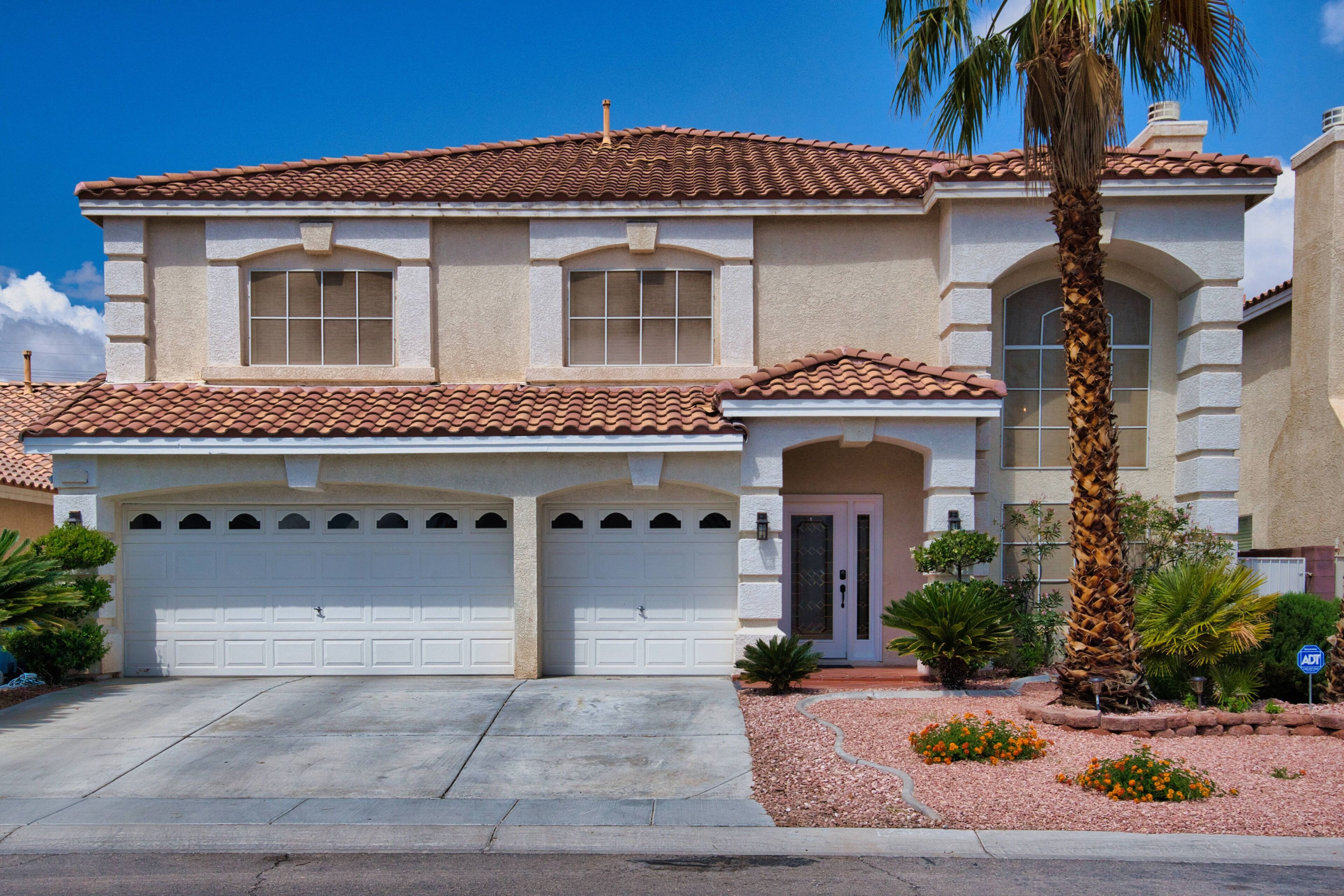 A two-story house with a tiled roof, a garage, and a well-maintained garden with palm trees and flowers.