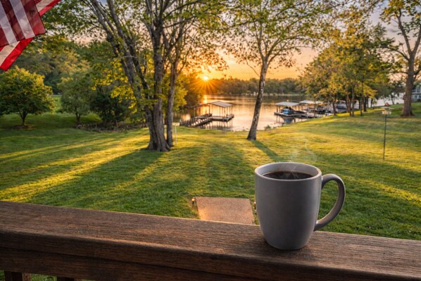 Une terrasse en bois avec une tasse de café, donnant sur un lac où l’on aperçoit des bateaux et un coucher de soleil.