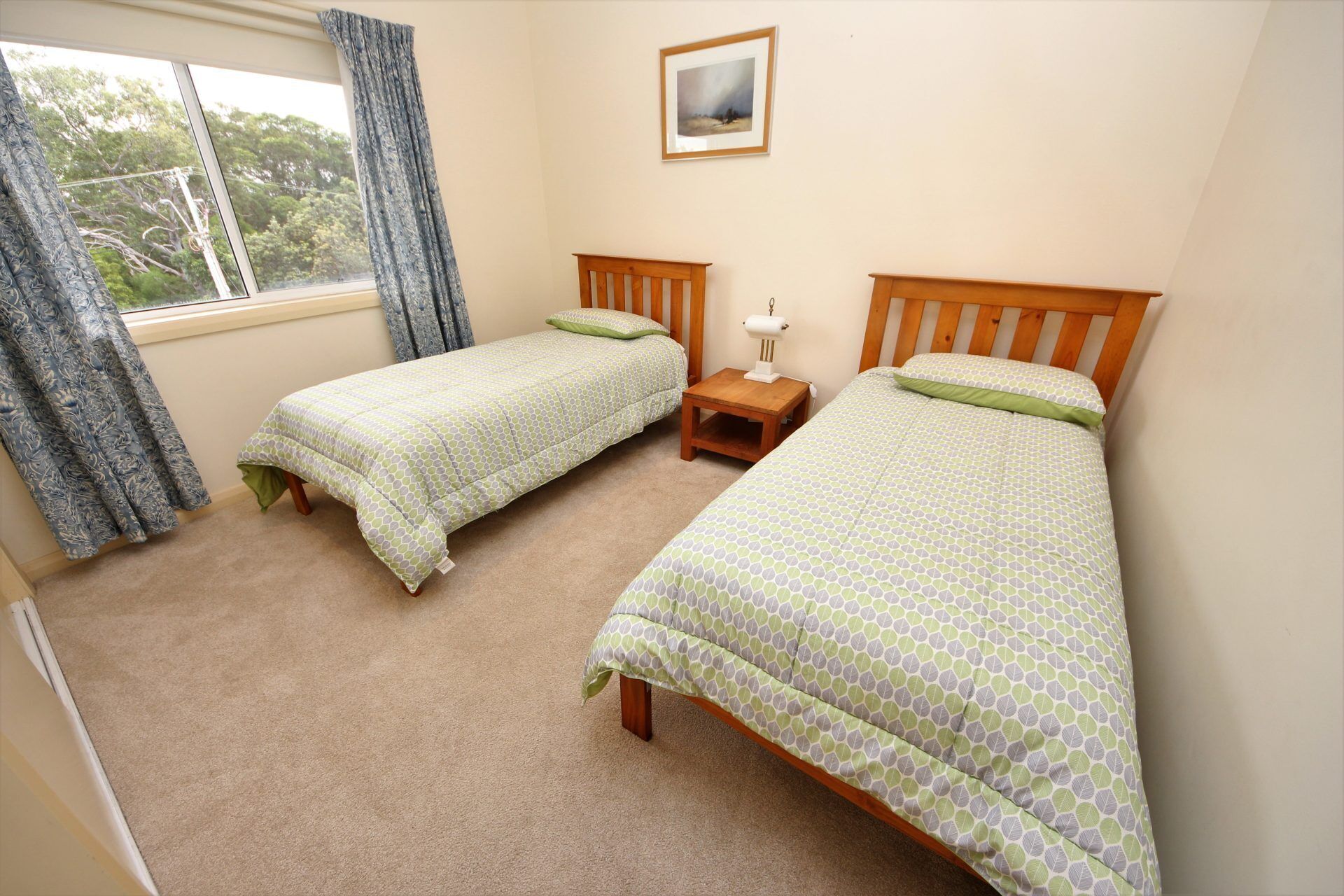 A room with two single beds, a small bedside table with a lamp, a window with curtains, and a framed picture on the wall.