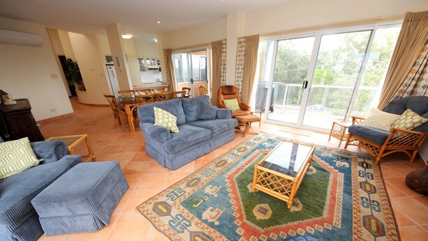 A spacious living room with a blue sofa, a patterned rug, and a dining area with a table and chairs. - Stunning Penthouse in the Heart of Town – Views, WiFi & Walk to Everything (South West Rocks)