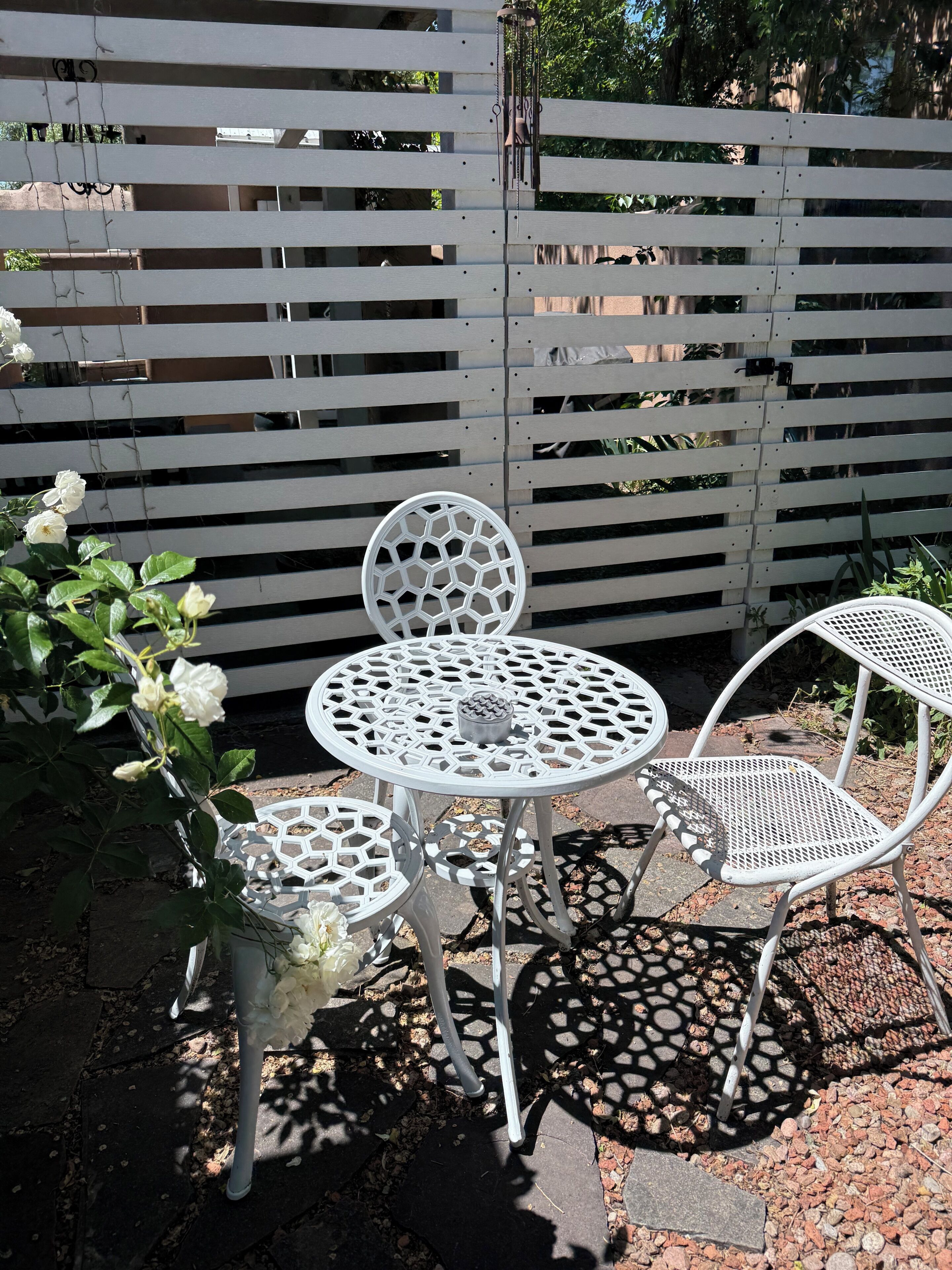 Et hvidt metalbord og to stole med et sekskantet mønster, placeret på en stenbelagt terrasse med et hvidt hegn i baggrunden.