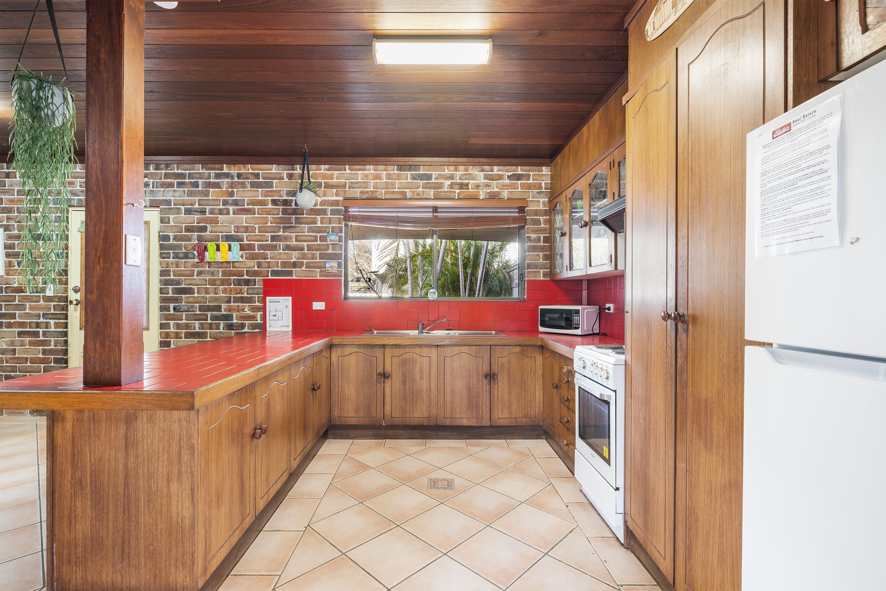 A kitchen with wooden cabinets, a red countertop, a brick wall, and a tiled floor.