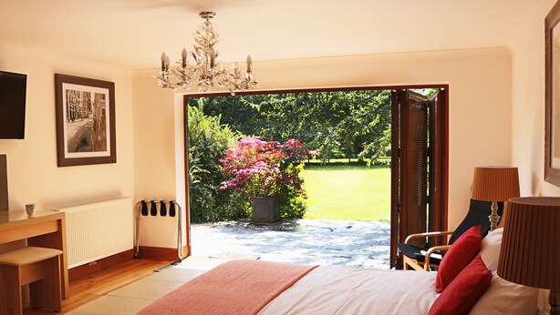 Ein Schlafzimmer mit einem Bett, einem Schreibtisch, einem Stuhl, einem Heizkörper, einer Chandelier und Blick auf einen Garten durch eine offene Tür.