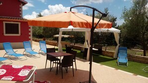 An outdoor patio with a table and chairs, a covered seating area, and a red building in the background. - Villa manghisi holiday home (Noto, Siracusa)