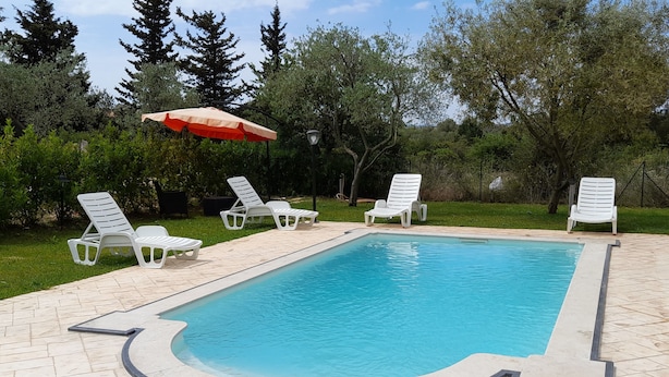 A rectangular swimming pool with a tiled deck, surrounded by white lounge chairs and a red umbrella. - Villa manghisi holiday home (Noto, Siracusa)