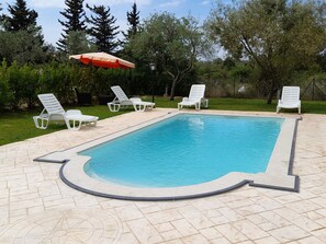 Ein rechteckiger Pool mit Fliesen umgeben, mit weißen Liegestühlen und einem roten Sonnenschirm.