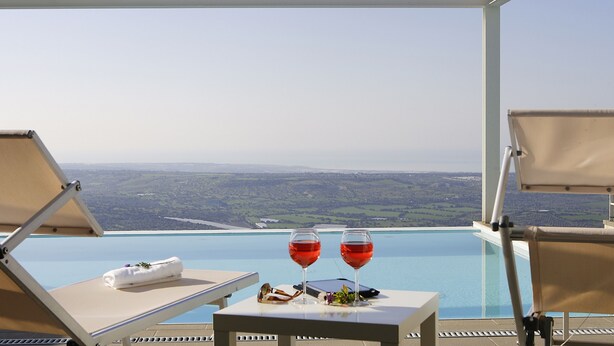 Ein Infinity-Pool mit Blick auf die Landschaft, zwei Liegestühle, ein Tisch mit Handtuch und Gläsern sowie zwei Gläser Rotwein.
