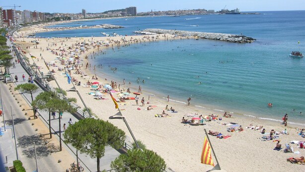 Een strand met mensen die zonnen en zwemmen, een rotsenrijke zeewering en gebouwen op de achtergrond.