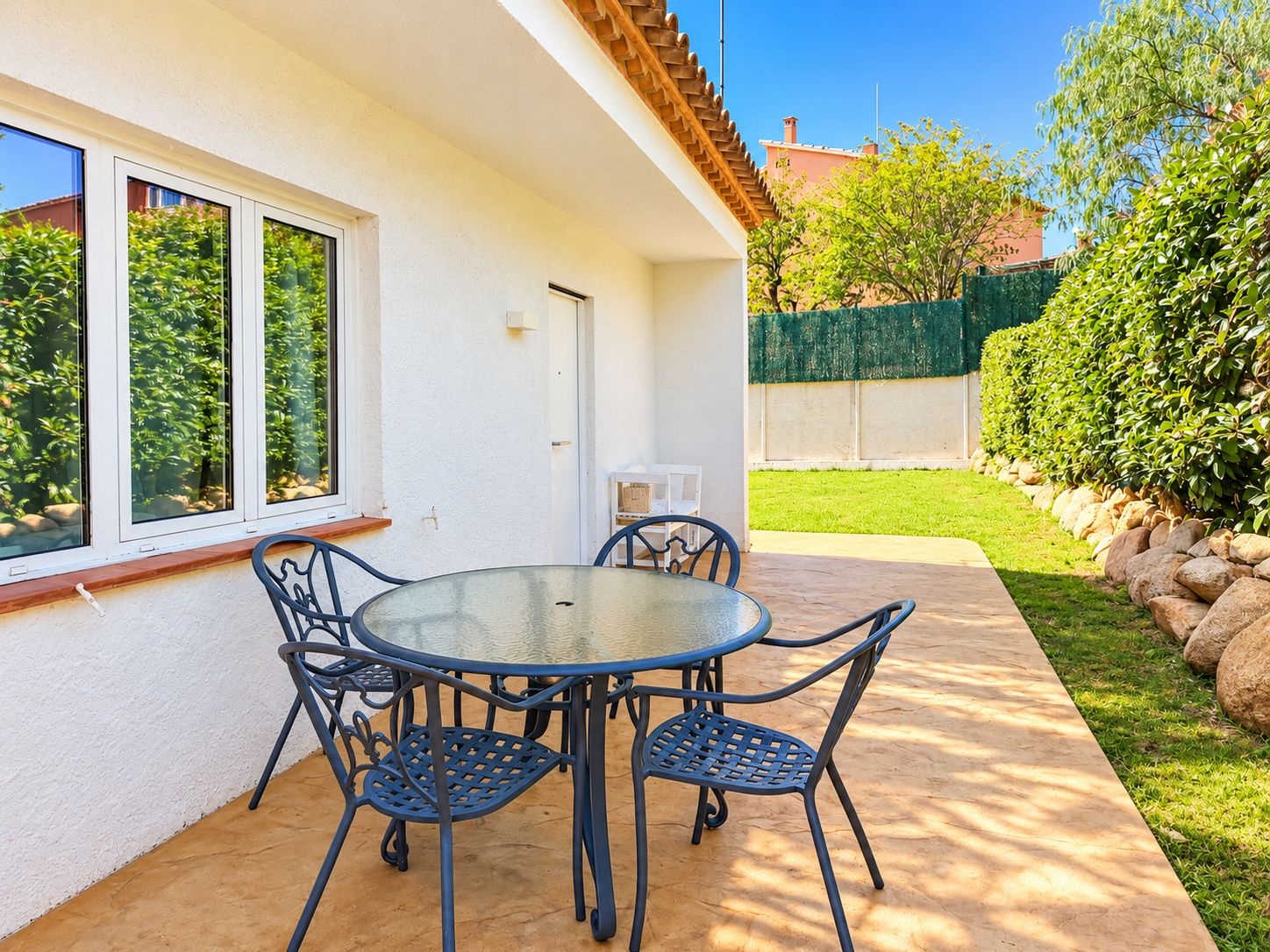 A patio with a round table and chairs, a white wall, and a view of a green lawn and trees.