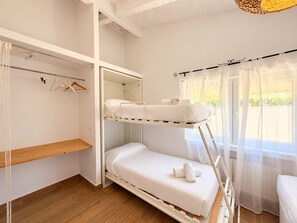 Une chambre avec un lit superposé, une étagère en bois, un porte-vêtements et une fenêtre avec des rideaux légers.