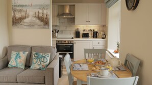A cozy living room with a sofa and a painting, a wooden dining table with breakfast items, a kitchen with white cabinets and modern appliances. - Apartment set in stunning grounds with coastal views & access to coastal paths (Thurlestone)