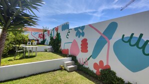 Un patio con pared blanca decorada con arte abstracto de colores, una pequeña mesa con sillas y una palmera.