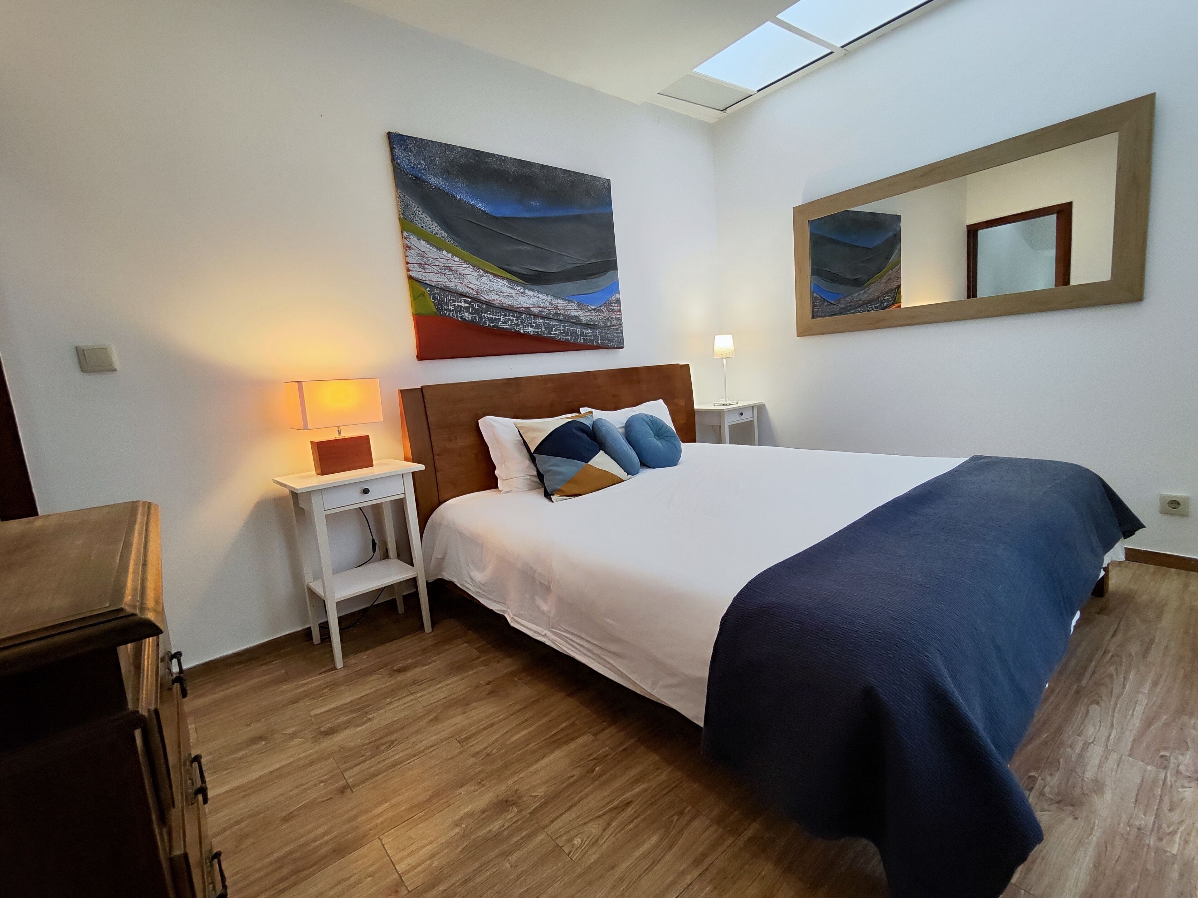 A hotel room with a large bed, a nightstand with a lamp, a painting on the wall, and a skylight above.