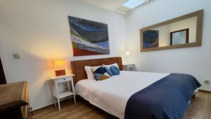 Habitación de hotel con una cama grande, una mesita de noche con una lámpara, un cuadro en la pared y un tragaluz arriba.