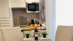 Ein Essbereich mit Korbstühlen und einem weißen Tisch, eine Küche mit Mikrowelle und Obstschale, sowie ein kleiner Topfpflanze auf dem Tisch.