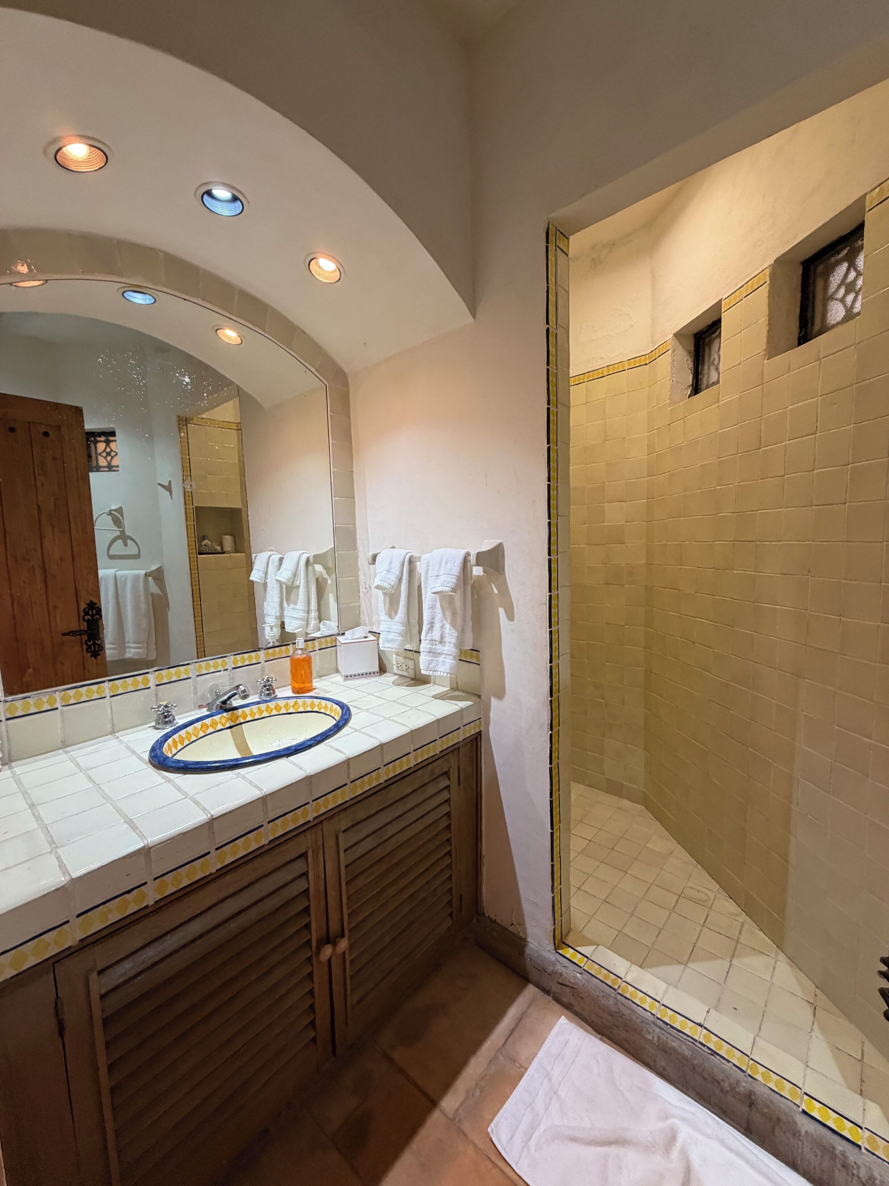 Un baño con un espejo grande, un lavamanos con borde azul, una ducha con puerta de vidrio y un tocador de madera.