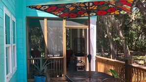 Una casa turquesa con una terraza de madera, una mesa y sillas de metal, un paraguas de colores y una planta en maceta.