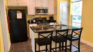Una cocina con armarios blancos, un refrigerador negro, un microondas, una estufa y una isla de vidrio con dos sillas.