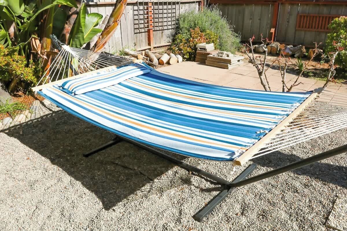 A hammock in a backyard with a wooden fence, gravel ground, and various plants.