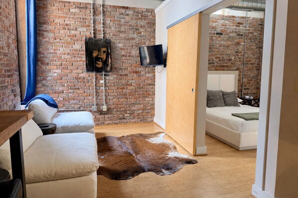 A modern room with a brick wall, a mounted TV, a white sofa, a leather rug, and a bed with patterned pillows. - Downtown Music City Loft living! ~Steps to the Ryman and Broadway (Nashville)