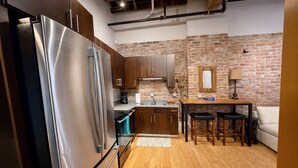A modern kitchen with stainless steel appliances, wooden cabinets, a brick wall, and a cozy seating area with a table and chairs. - Downtown Music City Loft living! ~Steps to the Ryman and Broadway (Nashville)