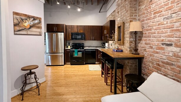 A compact kitchen with a refrigerator, microwave, and stove, wooden flooring, and a brick wall. - Downtown Music City Loft living! ~Steps to the Ryman and Broadway (Nashville)