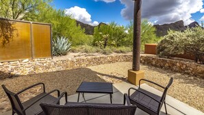 Une terrasse extérieure avec deux fauteuils en osier et une petite table, entourée d’un mur de pierre et d’une végétation désertique, avec des montagnes en arrière-plan.