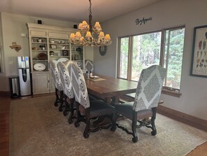 A dining area with a chandelier, patterned chairs, a wooden table, and a window with a view of trees. - Large, luxury cabin, sleeps 19 with a gourmet kitchen, hot tub & fire pit (Angel Fire)